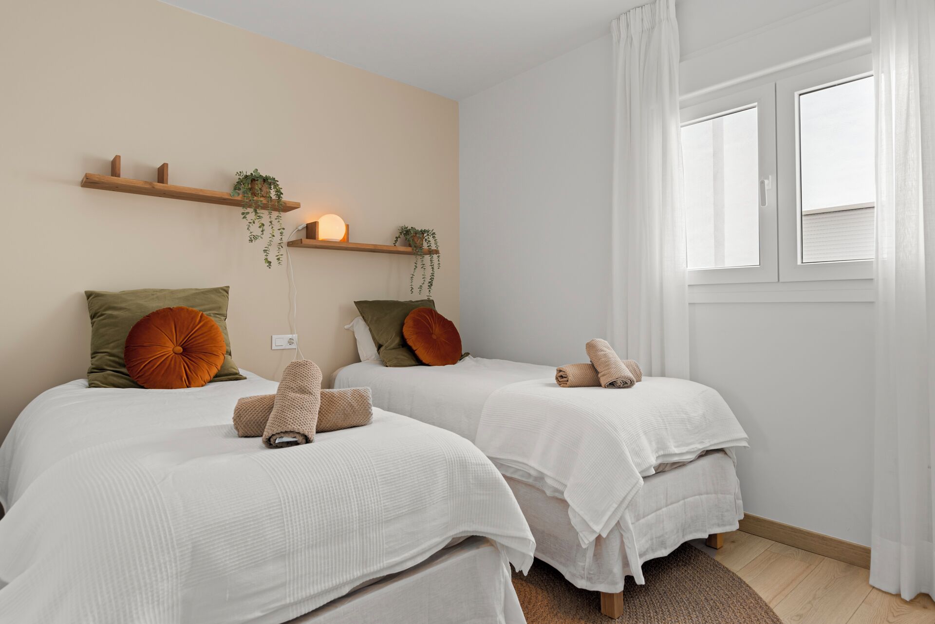 A bedroom with two beds, white bedding, green and orange pillows, a hanging plant, and a window with white curtains.