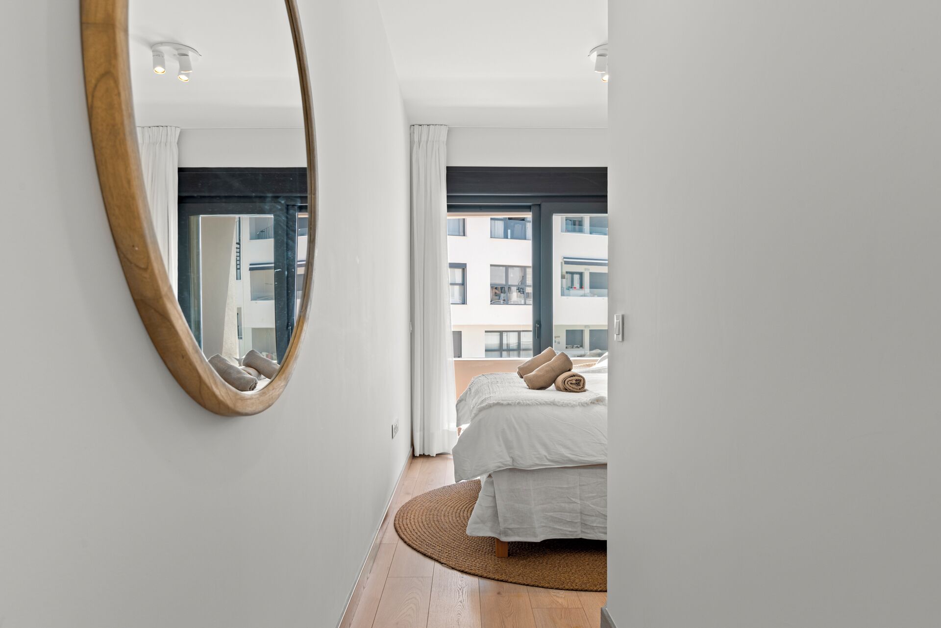 Une chambre à coucher avec un lit, un miroir rond et une vue sur un immeuble par la fenêtre.