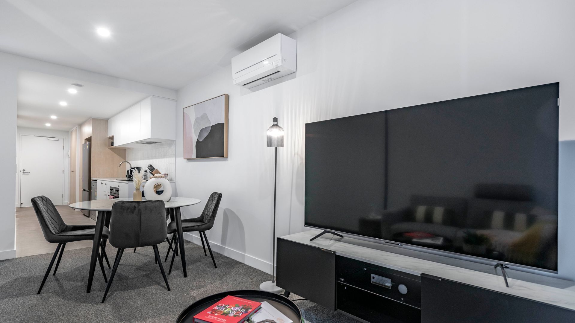 Een moderne woonkamer met een grote flatscreen televisie, een eethoek met een ronde tafel en stoelen, en een keuken op de achtergrond.