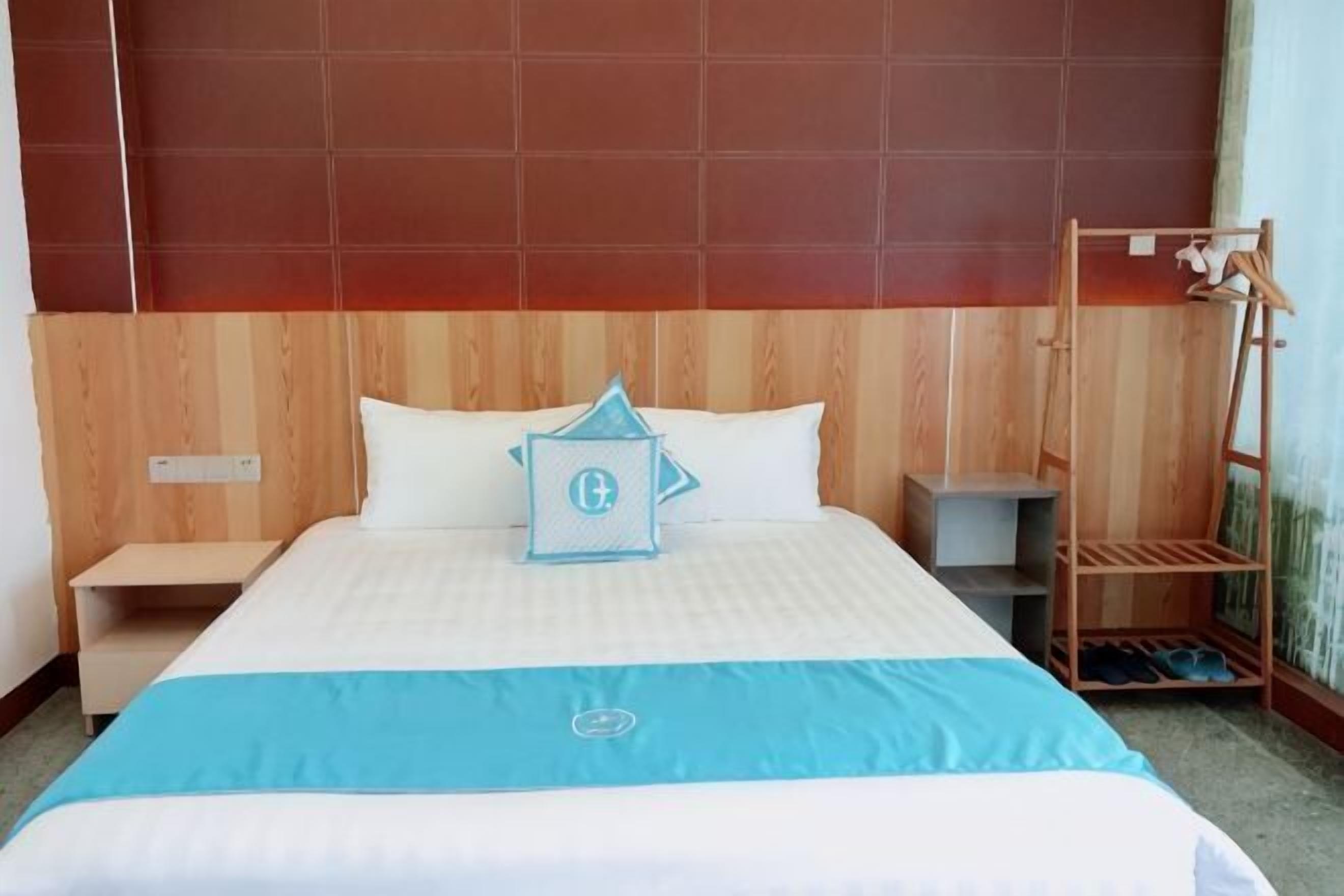Hotellihuone, jossa on puinen sängynpääty, sinivalkoinen peitto sängyllä, yöpöytä ja puinen hyllykkö.