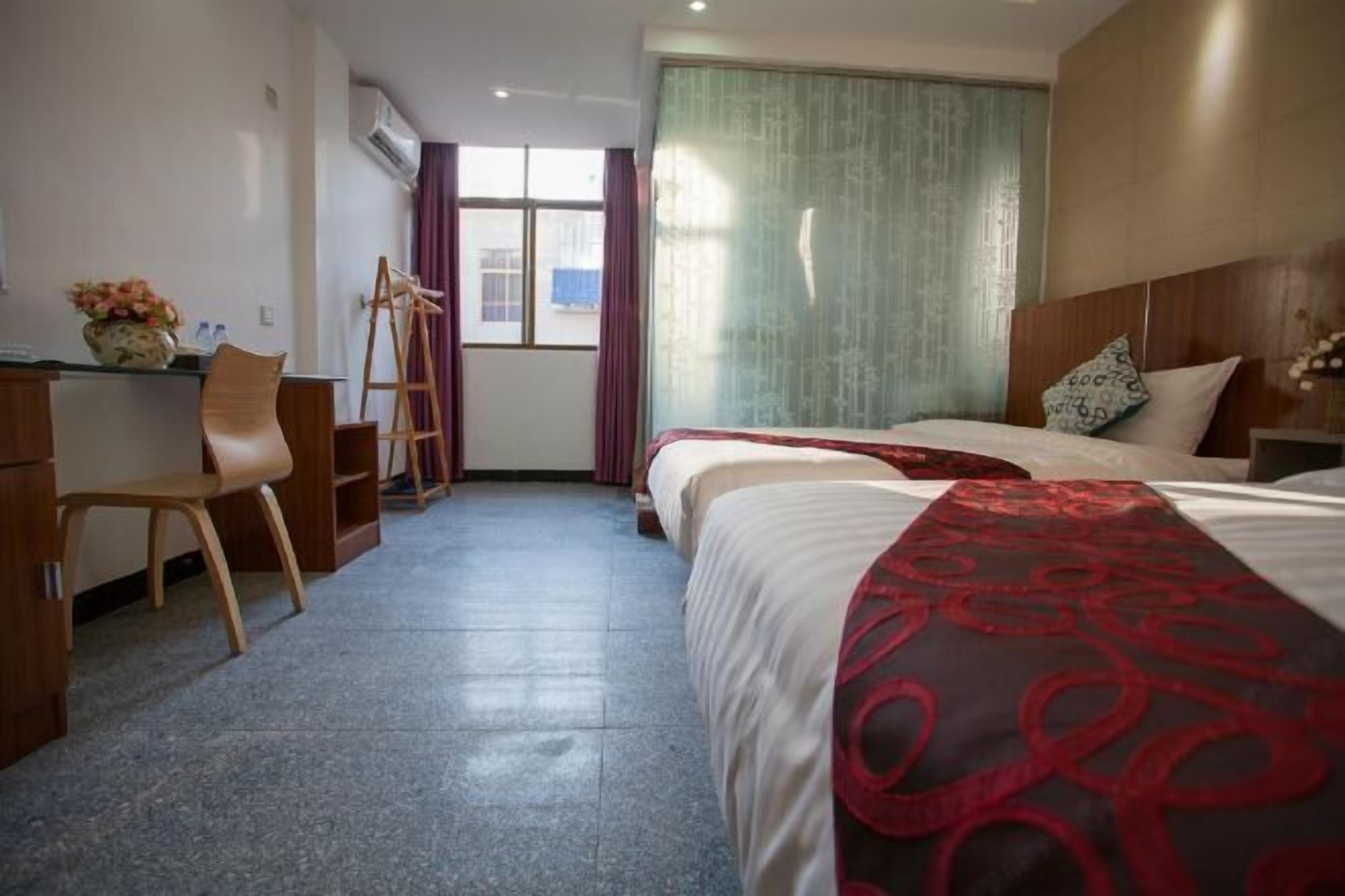 A hotel room with a desk, two beds, a chair, a ladder, and a window with curtains.