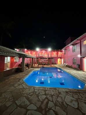 A courtyard with a swimming pool, stone flooring, and pink buildings with balconies and windows. - Pousada Villa Rosada  (Santa Cruz Cabralia)