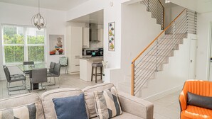 Ein modernes Wohnzimmer mit einer Couch, einem Esstisch und Stühlen. Eine Küchenzeile mit Herd und Schränken ist ebenfalls vorhanden. Eine Holztreppe führt auf eine weitere Ebene.