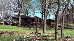 A rustic log cabin with a porch, surrounded by a well-maintained lawn and flowering plants. - Captains Cottage is the perfect retreat (Afton)