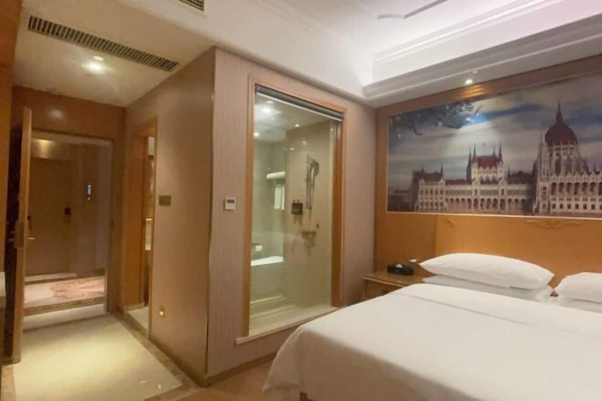 Una habitación de hotel con una cama, ducha con mampara de cristal, una mesita de noche y un cuadro de un edificio.