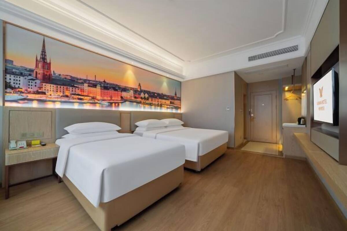 Habitación de hotel con dos camas, un cuadro de un paisaje urbano, un escritorio con lámpara y un televisor en la pared.