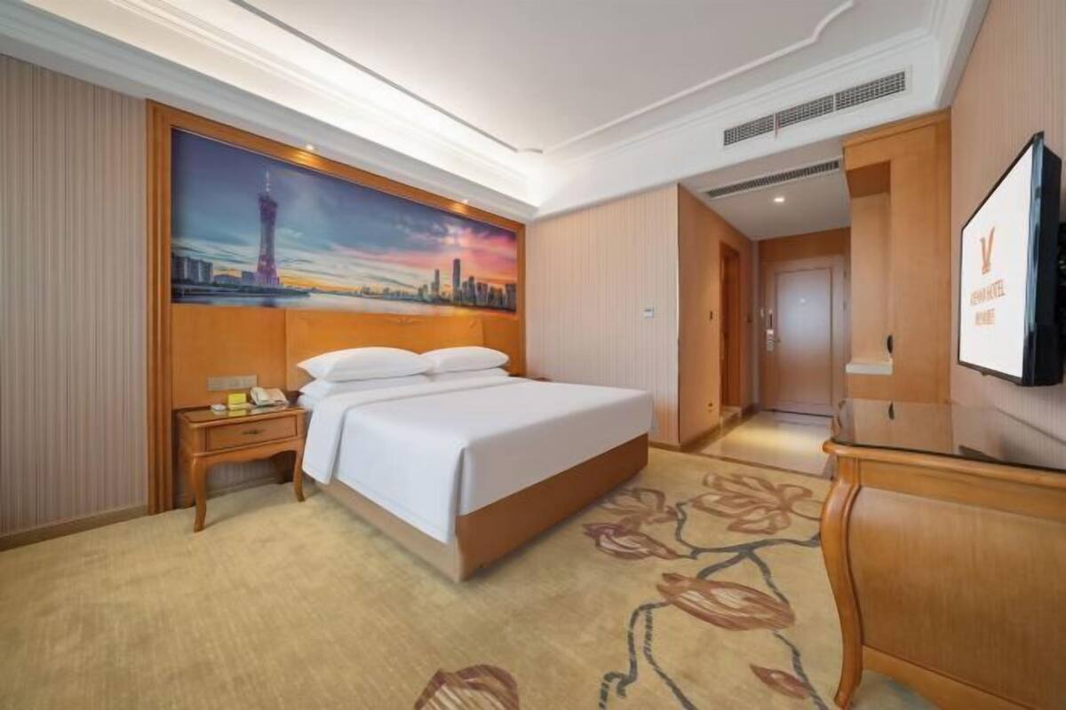 Una habitación de hotel con una cama grande, una mesita de noche, un televisor y un cuadro de un paisaje urbano.