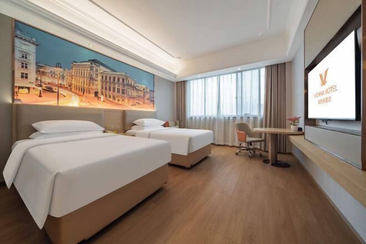 Habitación de hotel con dos camas, un escritorio, una silla, un televisor y un cuadro de un paisaje urbano en la pared.