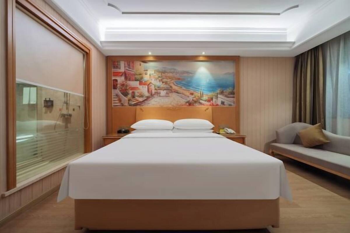 Habitación de hotel con una cama grande, un cuadro de una escena costera en la pared, un sofá y una ventana con persianas.