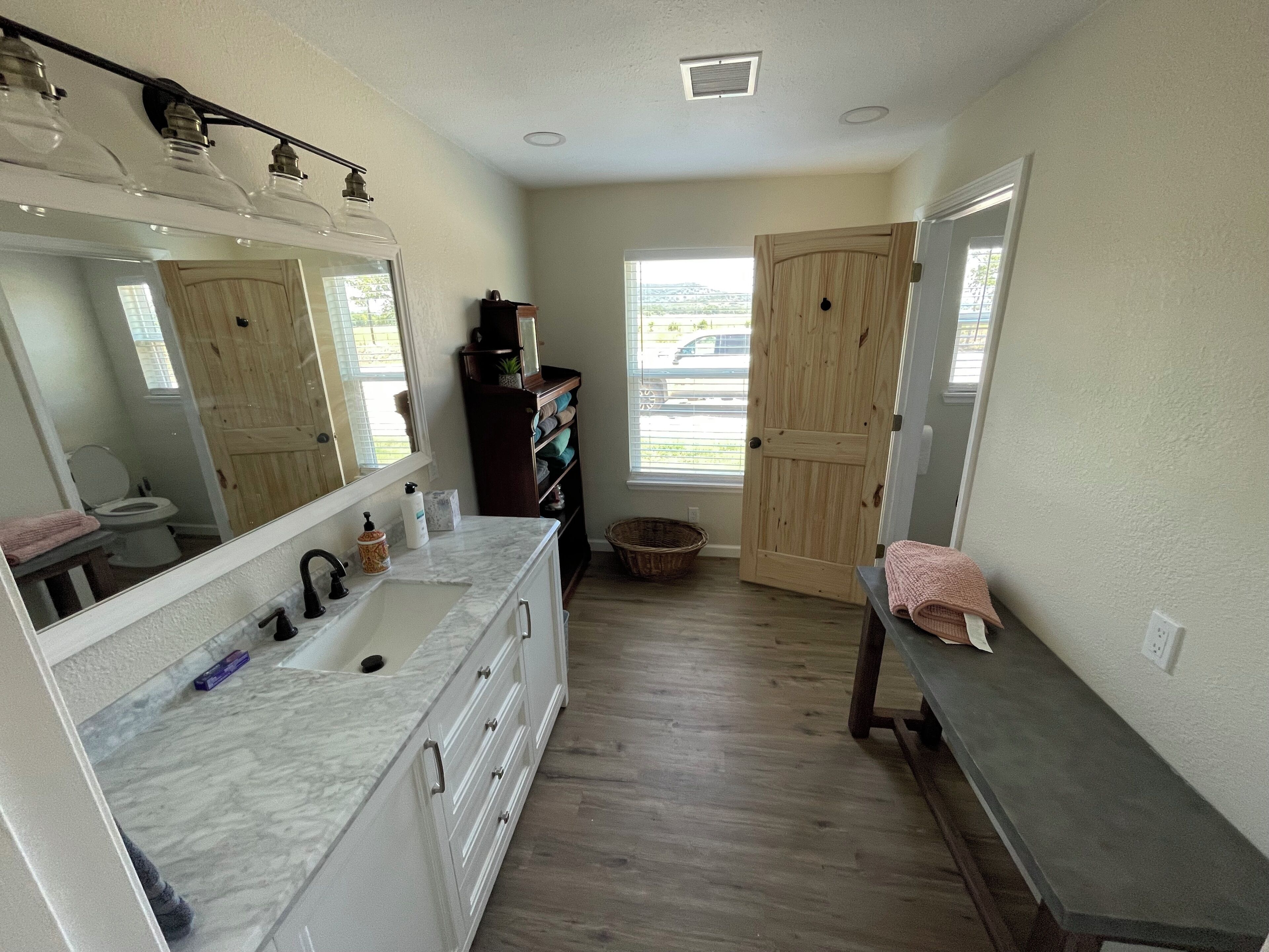 A bathroom with a large mirror, white cabinets, a marble countertop, wooden flooring, a wooden door, and a window with a view.
