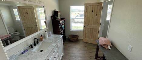 A bathroom with a large mirror, white cabinets, a marble countertop, wooden flooring, a wooden door, and a window with a view.