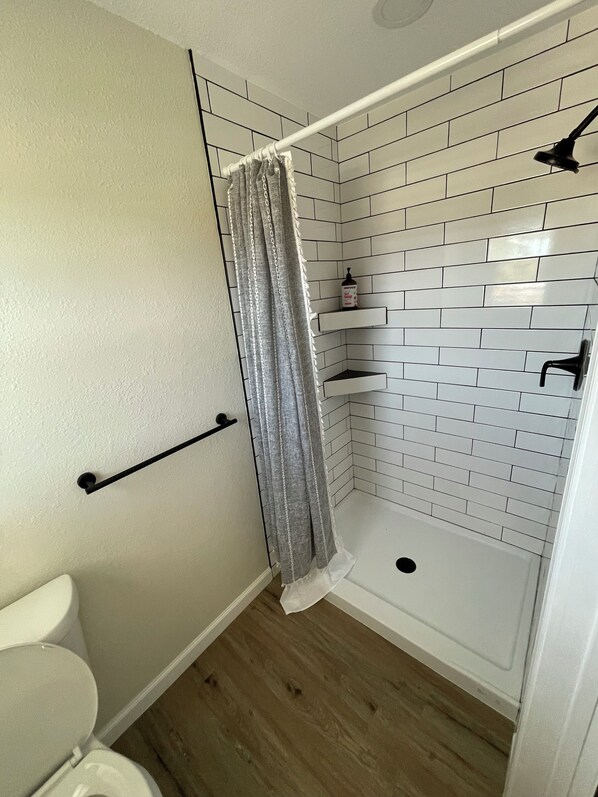 A bathroom with a white toilet, a shower with a white tiled wall, and a wooden floor. - Rio Verde - Cozy Cottage on the Frio River (Leakey)