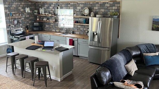 A modern kitchen with a central island, stainless steel appliances, and a dining area with a black leather couch. - Rio Verde - Cozy Cottage on the Frio River  (Leakey)
