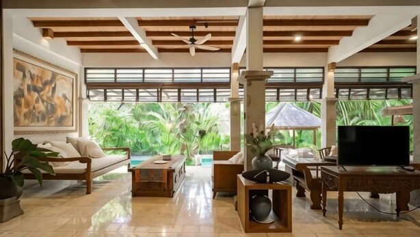 A spacious living area with a sofa, coffee table, TV, and large windows offering a view of greenery. - The Lavana Ziza Living And Villas Seminyak (Bali)