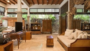 A spacious living area with a sofa, TV, wooden furniture, and large windows with a view of greenery. - The Lavana Ziza Living And Villas Seminyak (Bali)