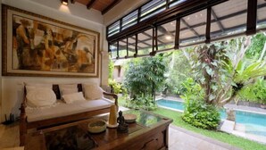 A room with a wooden sofa, a glass coffee table, and a large painting above it. There is a view of a swimming pool and lush greenery outside. - The Lavana Ziza Living And Villas Seminyak (Bali)