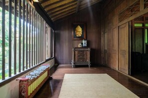 Un couloir doté d’un plancher en bois, d’une grande fenêtre avec des stores verticaux, d’une statue de Bouddha et d’un meuble traditionnel.