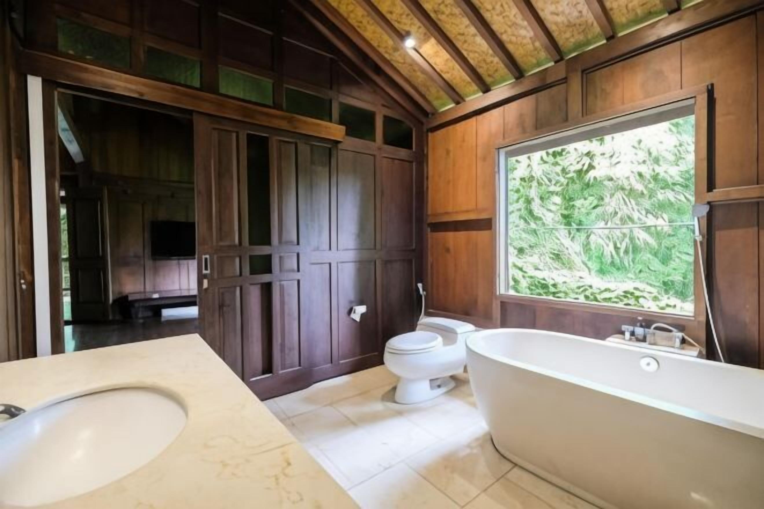 Un baño con una bañera grande, inodoro y lavamanos, paneles de madera y ventana con vista a la vegetación.