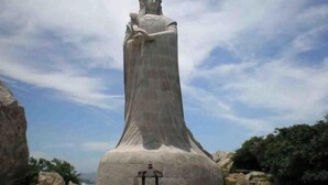 Uma grande estátua de pedra de uma mulher com manto esvoaçante, em uma plataforma com um candeeiro à frente.