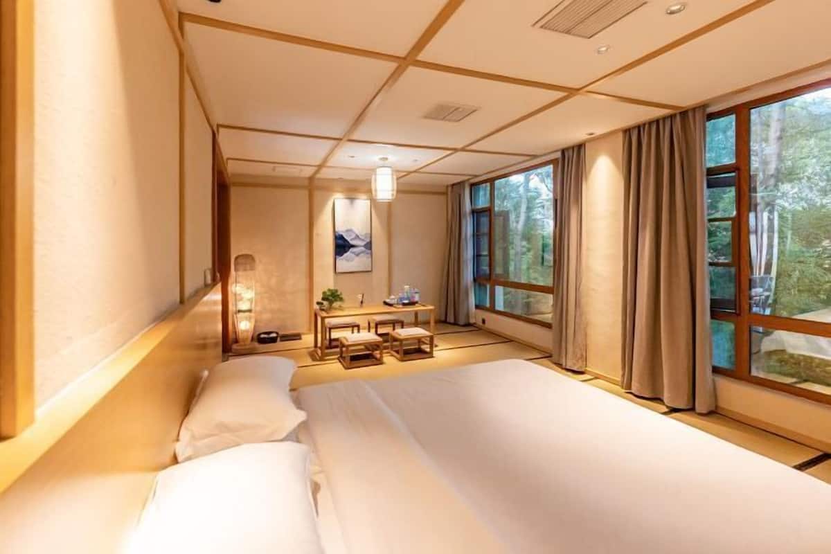 A hotel room with a large bed, a desk with a chair, a small table, and a window with curtains.