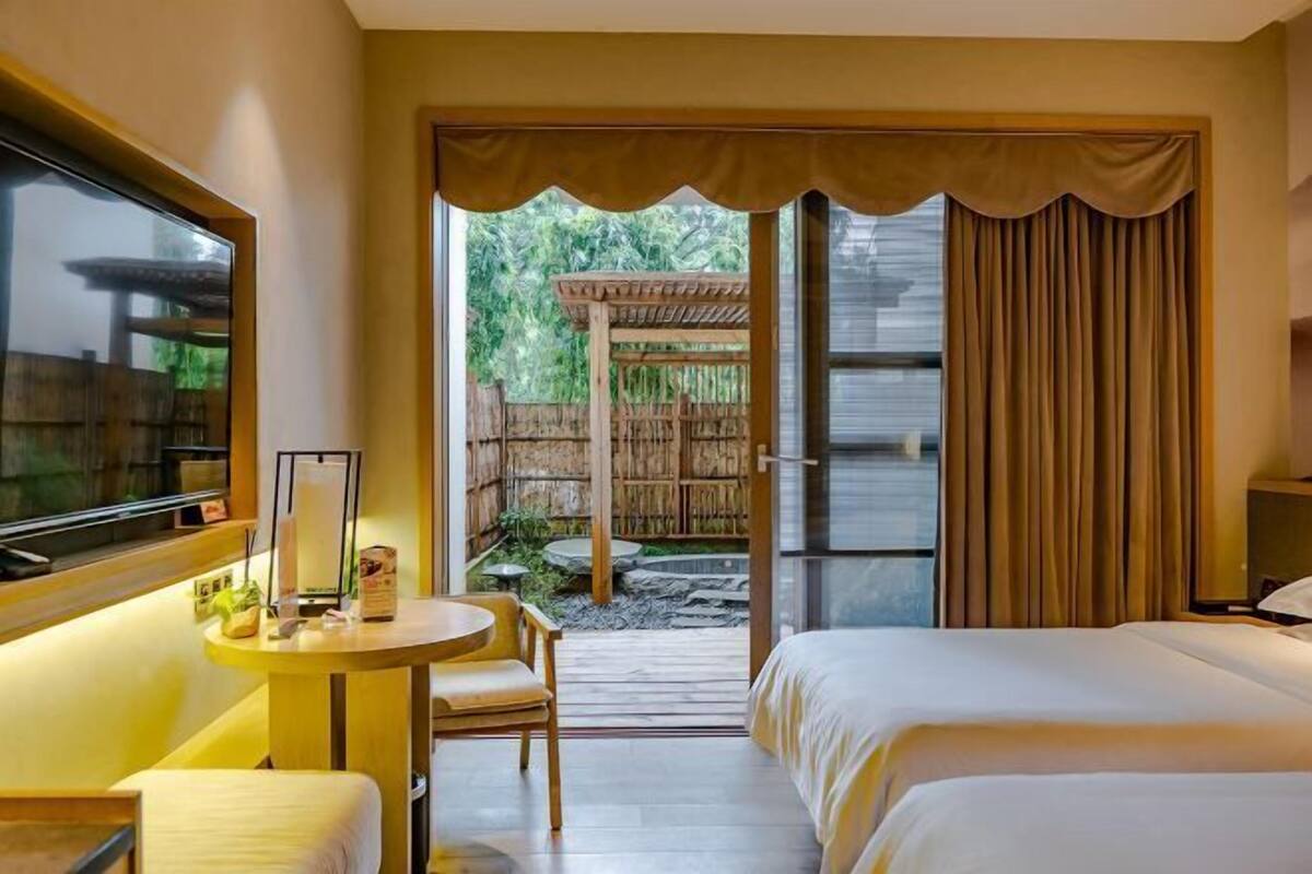 A hotel room with a bed, a desk with a chair, a window, and a view of an outdoor area with a wooden structure and greenery.