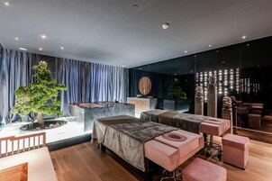 Una habitación de hotel moderna con una cama grande, dos mesitas de noche, una zona de estar con un taburete rosa y un amplio ventanal con cortinas.