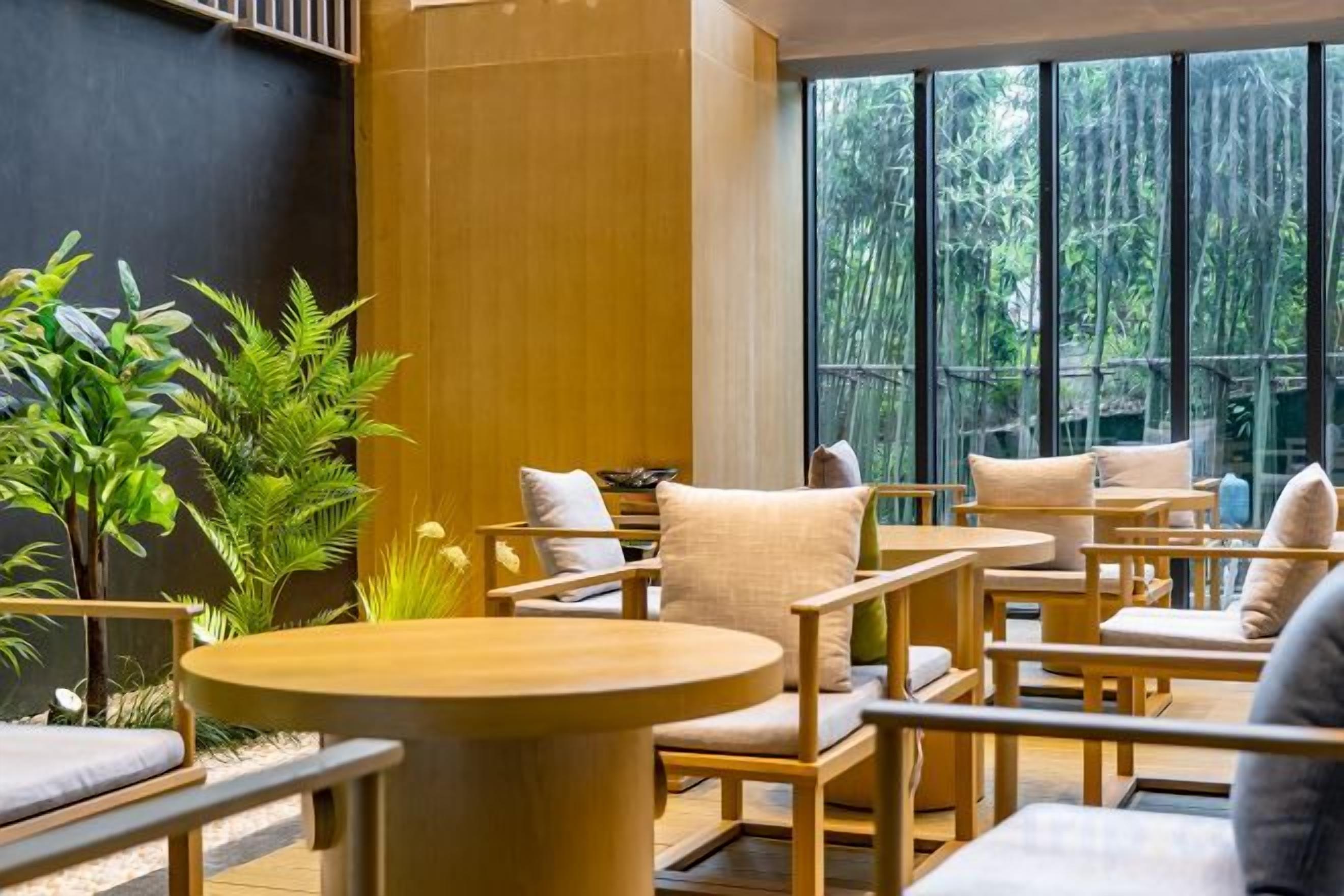 Un espacio interior moderno con asientos, mesas y sillas redondas, rodeado de ventanas amplias y plantas de interior.