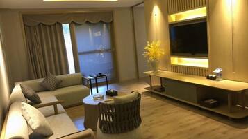 A modern living room with a sofa, ottoman, and a coffee table. There is a TV mounted on the wall, a vase with flowers, and a window with curtains.
