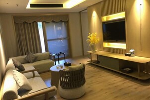 Una sala de estar moderna con sofá, reposapiés y una mesa de centro. Hay un televisor montado en la pared, un florero con flores y una ventana con cortinas.