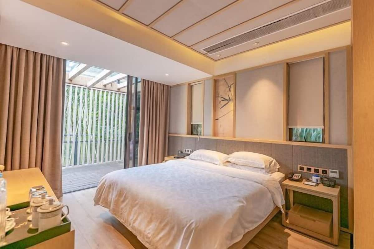 A modern hotel room with a bed, bedside tables, a desk with a chair, and a large window with curtains.