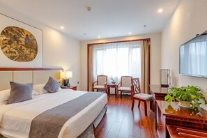 Une chambre d’hôtel avec un grand lit, deux chaises, une petite table, un téléviseur et un tableau au mur.