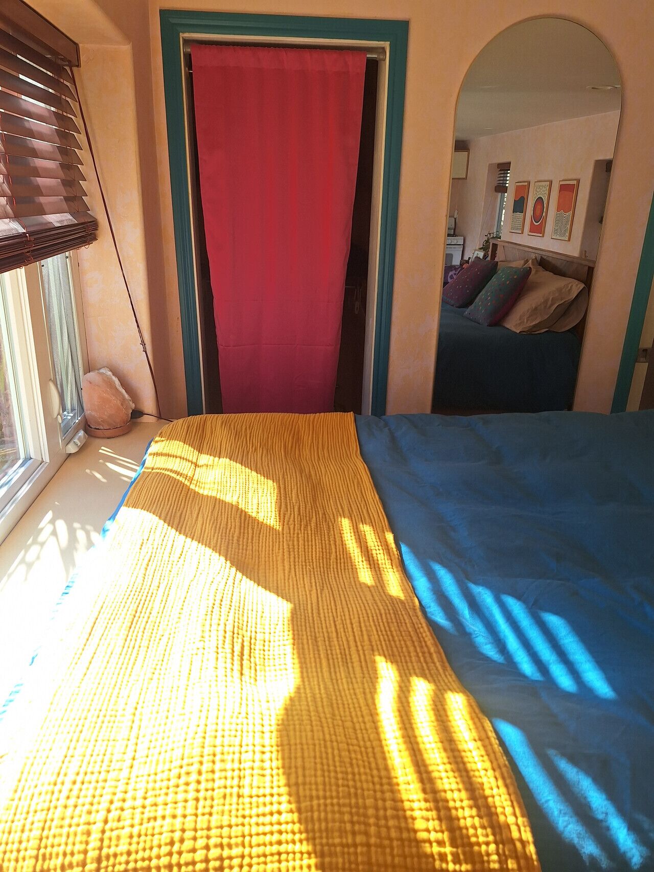 Een slaapkamer met een bed, een raam met zonwering, een rood gordijn en een spiegel die een andere kamer weerspiegelt.