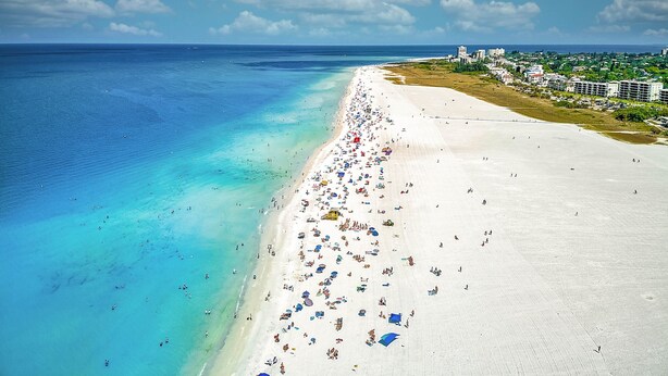 Una playa con una extensa franja de arena blanca, agua turquesa cristalina y algunos edificios al fondo.