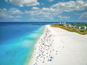 Une plage avec une longue étendue de sable blanc, une eau turquoise et limpide, et quelques bâtiments visibles au loin.