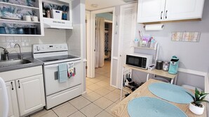 Cocina compacta con muebles blancos, estufa y microondas. Hay una zona de comedor con mesa y sillas.