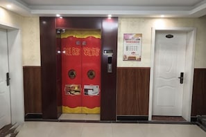 Entrada de hotel con un ascensor rojo y amarillo, una puerta blanca y un cartel en la pared.