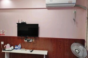 A room with a television mounted on the wall, a white table with a remote control, and a standing fan.