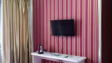 A room with a television mounted on a wall with striped wallpaper, a white console table with a kettle and a remote control, and a window with a curtain.