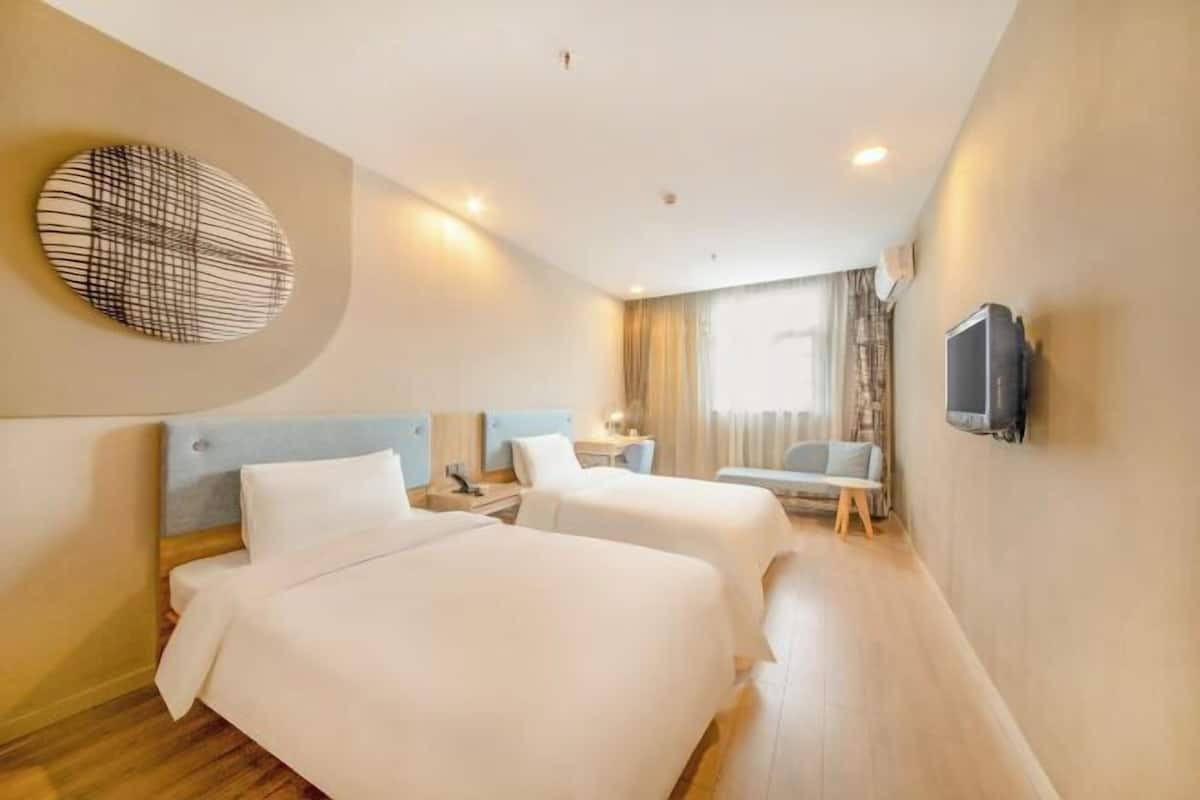 A hotel room with two beds, a TV mounted on the wall, a bedside table, and a window with curtains.