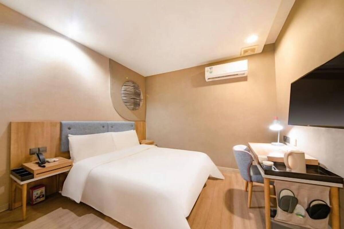 A bedroom with a bed, bedside table, desk, chair, and an air conditioning unit.