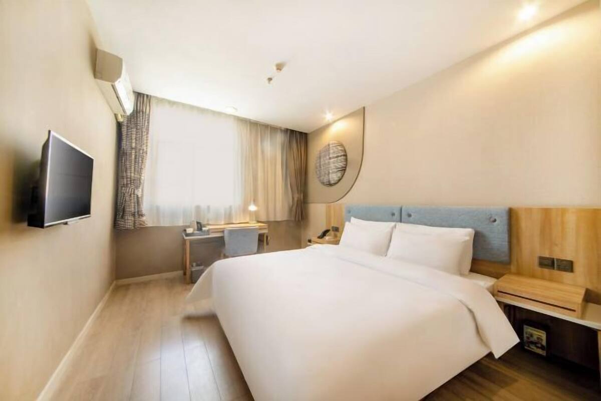 A hotel room with a bed, bedside table, lamp, TV, and a window with curtains.