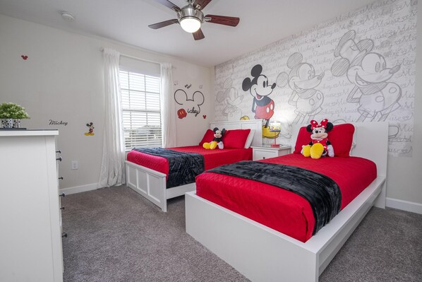 A bedroom with two beds, a ceiling fan, a window with curtains, and Mickey Mouse-themed decorations.