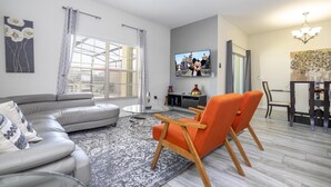 A modern living room with a grey sectional sofa, an orange armchair, a dining area with a table and chairs, a flat-screen TV, and a ceiling fan.