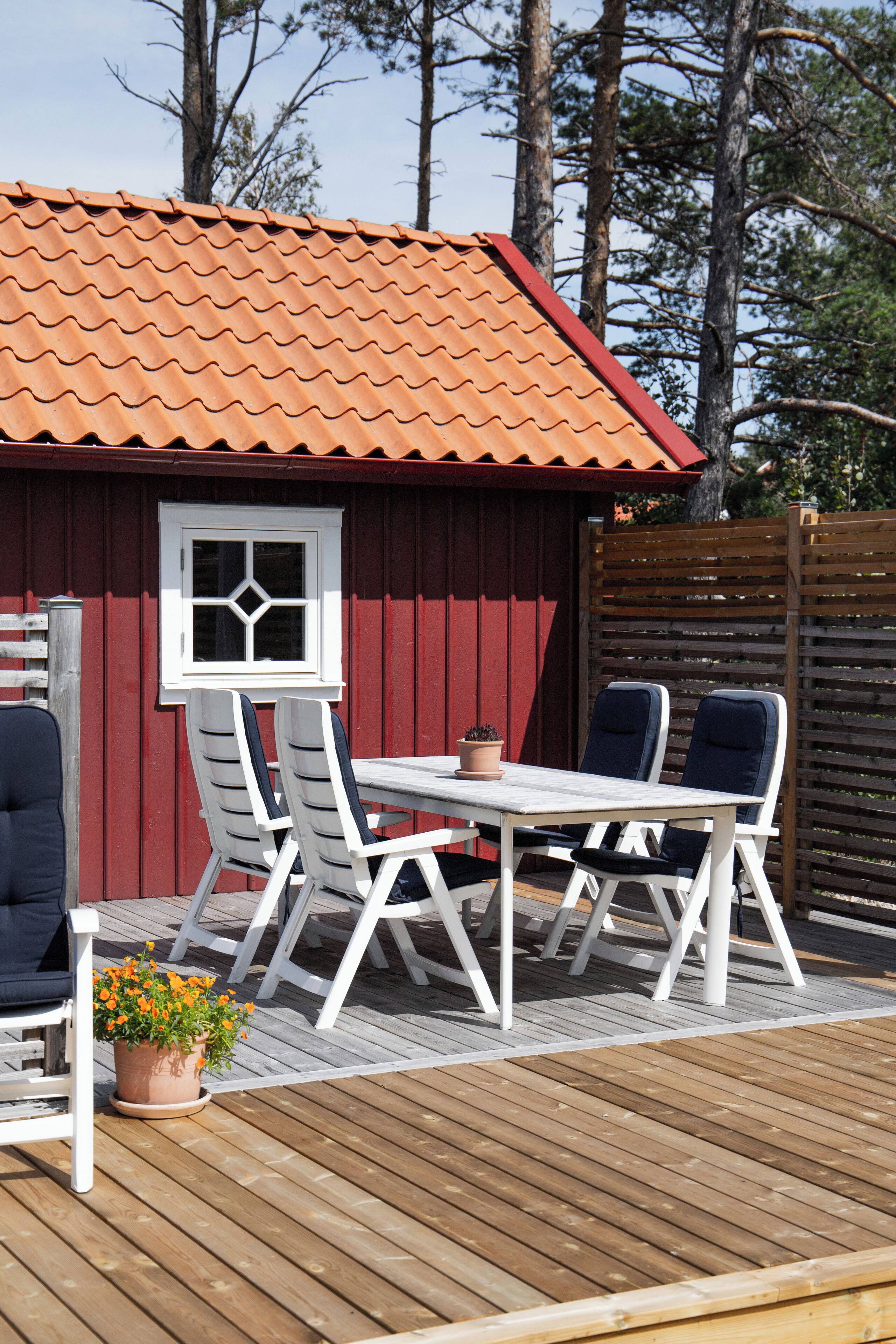 A wooden deck with a red building in the background, a white table with chairs, and a potted plant.