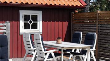 A wooden deck with a red building in the background, a white table with chairs, and a potted plant.