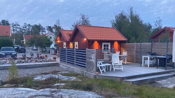 A wooden cabin with a red roof, a fenced patio area with outdoor seating, and a rocky foreground. - Charming 2-bedroom house new built in wonderful Grebbestad  (Grebbestad)