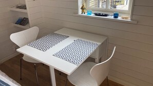 A small, white-framed table with a patterned tablecloth, white chairs, a window with a view of the outside, and a shelf with books and a small figurine. - Charming 2-bedroom house new built in wonderful Grebbestad  (Grebbestad)