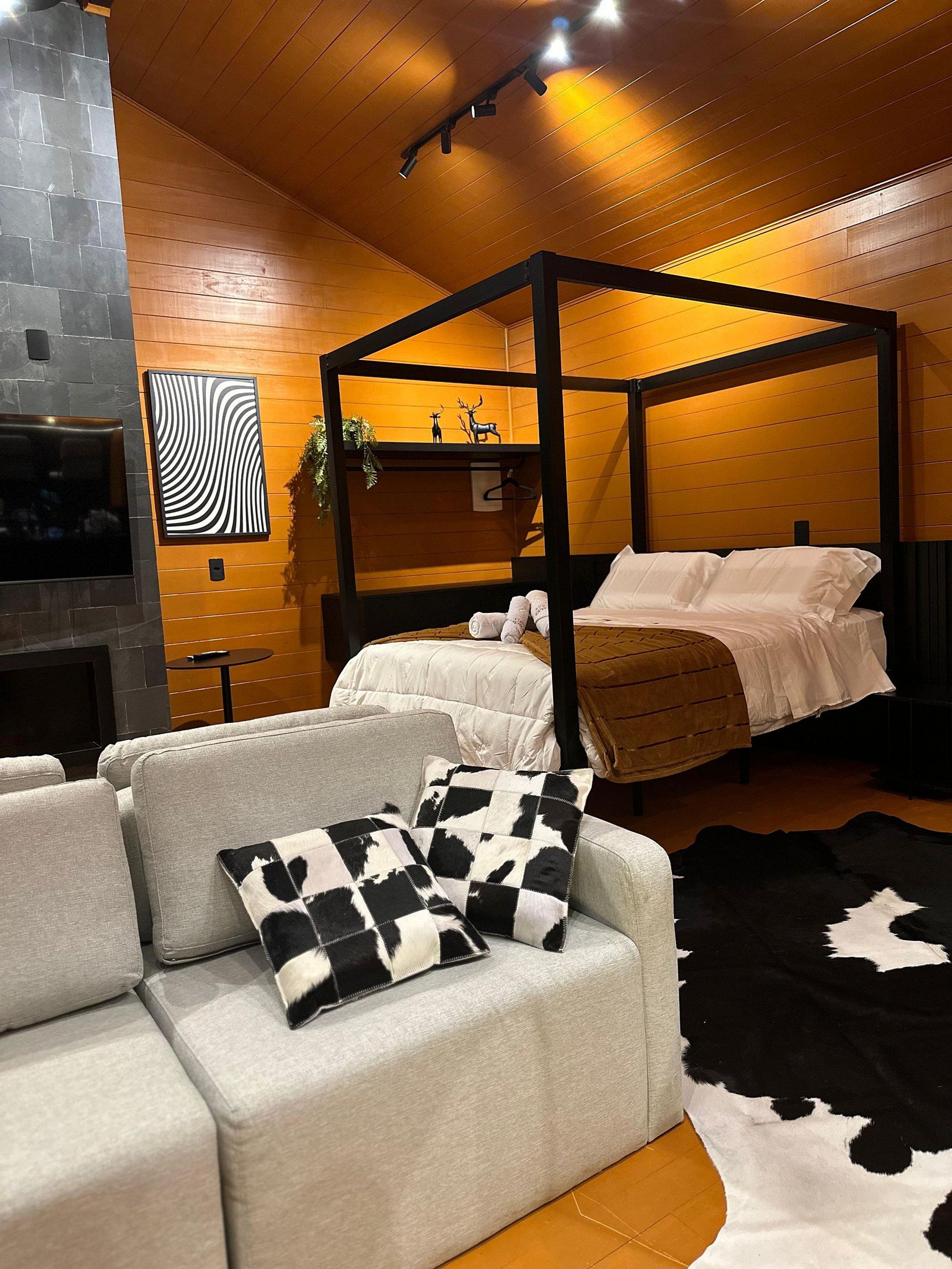 Een moderne slaapkamer met een hemelbed, een grijze bank met gekleurde kussens, een zwart-wit koeienhuidkleed en een aan de muur bevestigde televisie.