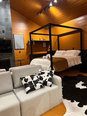 Un dormitorio moderno con una cama con dosel, un sofá gris con cojines estampados, un tapete de cuero de vaca en blanco y negro, y un televisor montado en la pared.