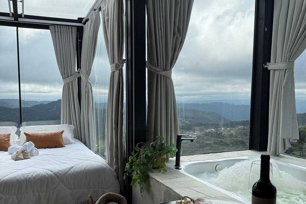 A bedroom with a bed, a view of the mountains, a bathtub, and a bottle of wine on a table.